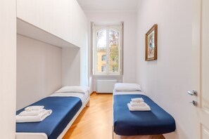 A narrow hotel room with two beds, a window with a view of buildings, and a framed picture on the wall. - Contemporary Flat at Trastevere (Roma)