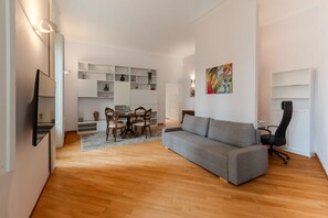 A modern living room with a grey sofa, wooden flooring, a dining area with a table and chairs, and a kitchen area with white cabinetry and shelves. - Contemporary Flat at Trastevere (Roma)