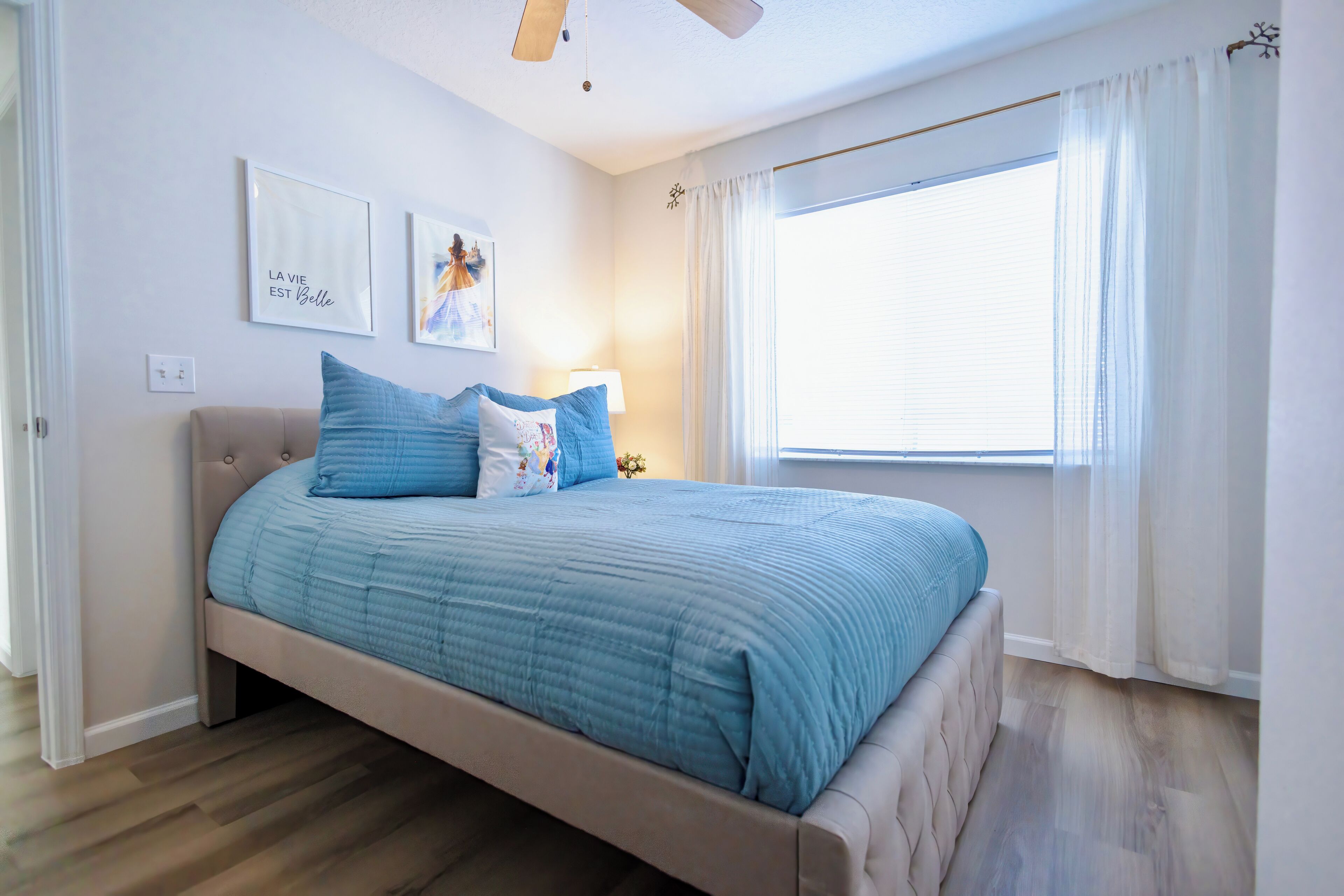 A bedroom with a bed, a ceiling fan, a window with curtains, and wall art.