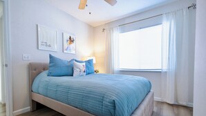 Une chambre à coucher comprenant un lit, un ventilateur de plafond, une fenêtre avec des rideaux et des œuvres d’art murales.