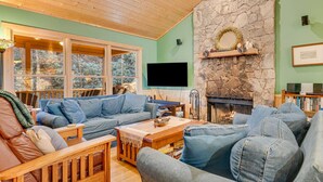 A cozy living room with a stone fireplace, wooden furniture, and a large window overlooking a forest.