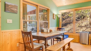 A wooden dining table with chairs, large windows, and a view of trees.