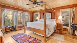 A wooden four-poster bed in a room with large windows, a ceiling fan, and a view of trees.