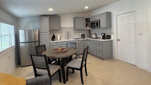 A modern kitchen with grey cabinets, a stainless steel refrigerator, a microwave, and a coffee maker. There is a dining table with chairs and a sofa in the foreground. - La Casita by Amarelys and lazaro (Jacksonville)