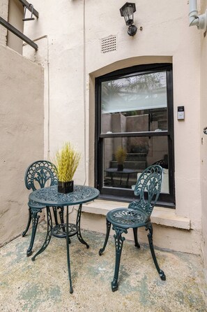 Un pequeño espacio al aire libre con dos sillas de metal y una mesa, una planta en maceta y una ventana con vistas a una escalera y vegetación.
