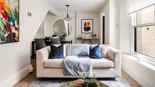 Ein modernes Wohnzimmer mit einem beigen Sofa, dekorativen Kissen, einem Couchtisch mit Blumen, einem großen Fenster und einem Essbereich im Hintergrund.