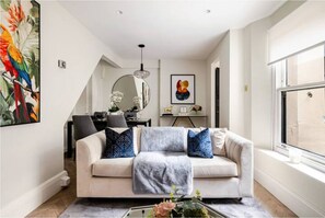 A modern living room with a beige sofa, decorative pillows, a coffee table with flowers, a large window, and a dining area in the background. - Boutique 3 Bedroom Apartment (London)