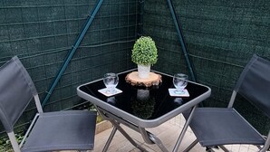 Een klein terras buiten met een tafel, twee stoelen, een decoratieve plant en een groene omheining.