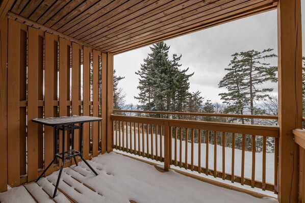A wooden balcony with a metal table, snow-covered ground, and a view of snow-covered trees.