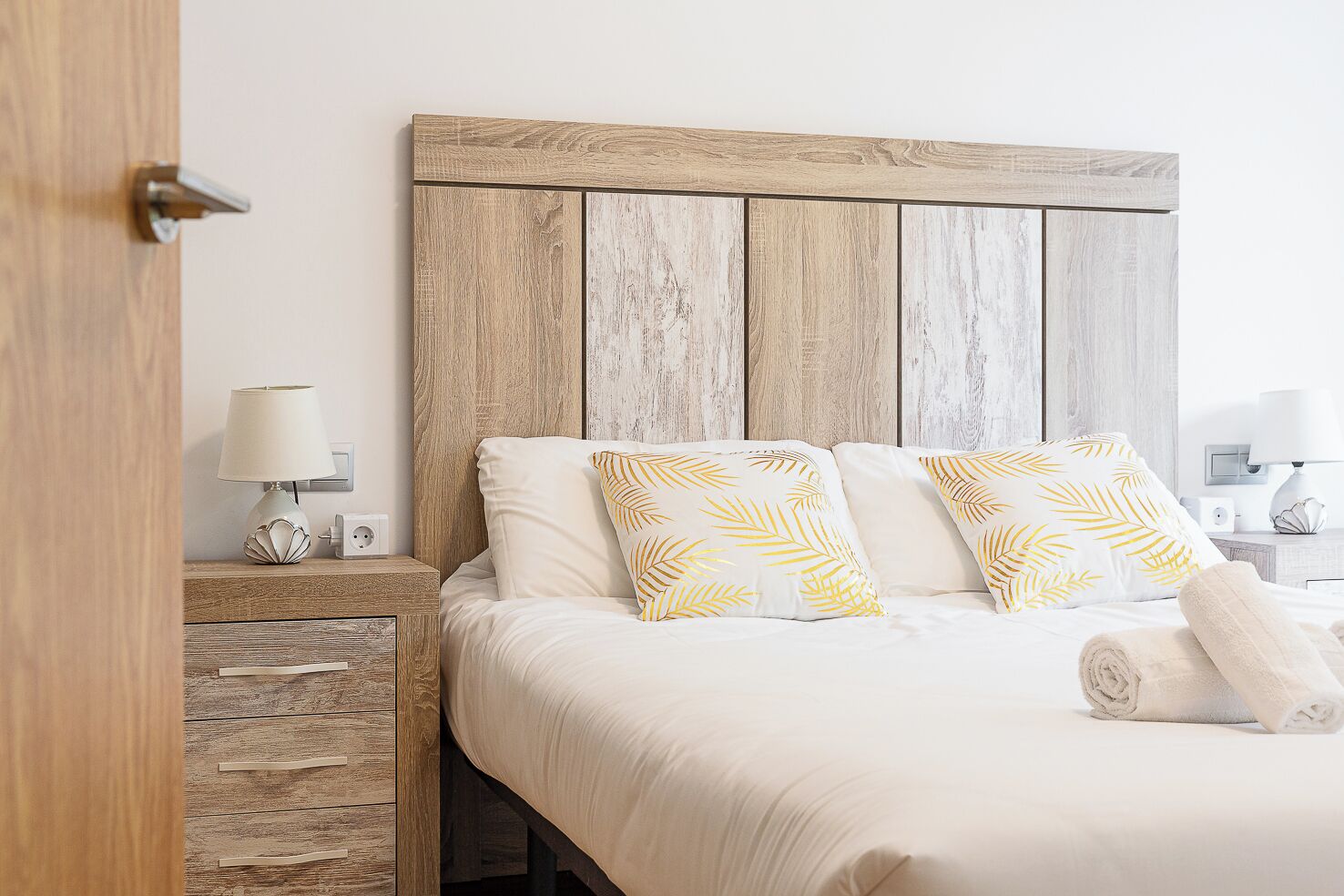 Un lit bien fait, avec une literie blanche et deux oreillers ornés d’un motif de feuilles, une tête de lit en bois, une table de chevet avec une lampe et une serviette enroulée posée sur le lit.