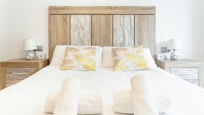 Un lit bien fait, avec du linge de lit blanc et deux oreillers ornés d’un motif de feuilles, flanqué de deux tables de chevet équipées de lampes et de prises électriques.