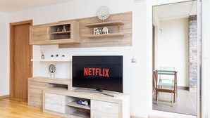 Un salon moderne avec un meuble TV en bois, un téléviseur à écran plat affichant le logo « NETFLIX », et une porte vitrée donnant sur un balcon.