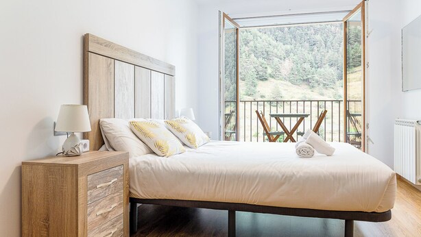 Une chambre avec un grand lit, une tête de lit en bois, une table de chevet et une vue sur une forêt, visible à travers une porte vitrée.