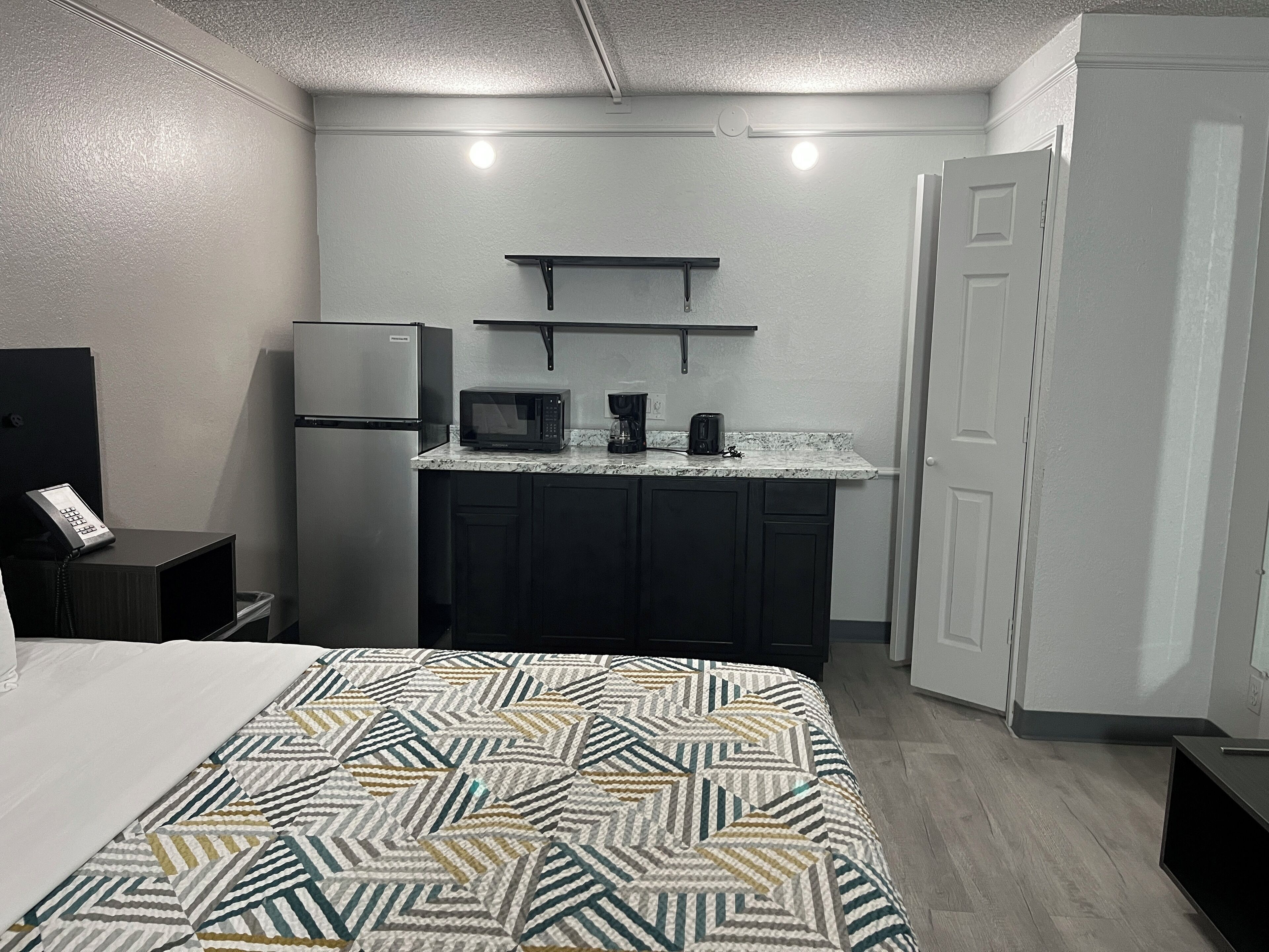 A compact hotel room with a bed, a kitchenette with a fridge, microwave, and coffee maker, and a door to the right.