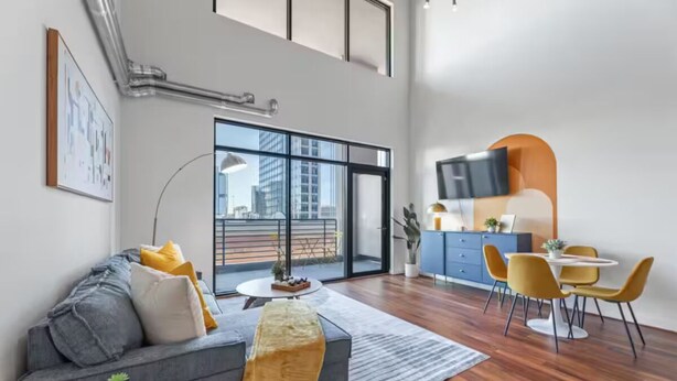Ein modernes Wohnzimmer mit grauem Sofa, gelben Kissen, einem runden Couchtisch und einem Essbereich mit gelben Stühlen und einem blauen Tisch.