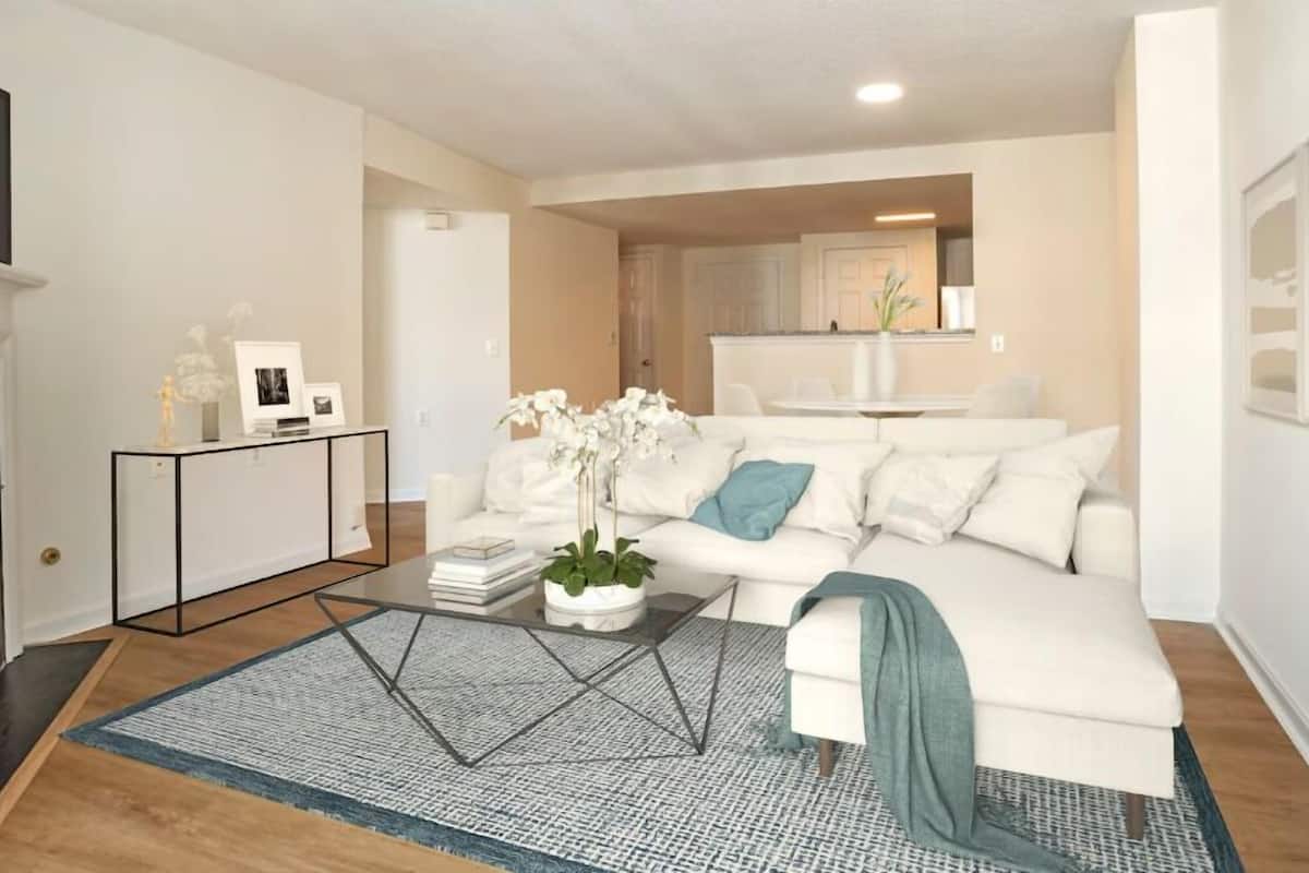 Un salon moderne avec un canapé blanc, une table basse sur laquelle sont posés des livres et un pot de fleurs, une cheminée, et une cuisine visible au fond.