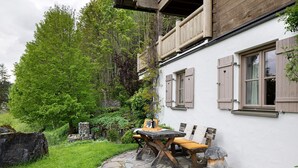 Eine Steinterrasse mit einem Holztisch und -stühlen, einer Steinmauer und einem weiß gestrichenen Gebäude mit Holzhinterfenstern.