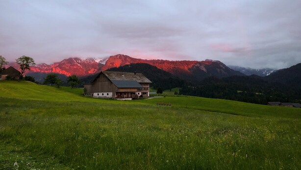 Ländliche Gegend mit einem einzelnen Haus auf einem grasbewachsen Hügel, umgeben von Bergen.