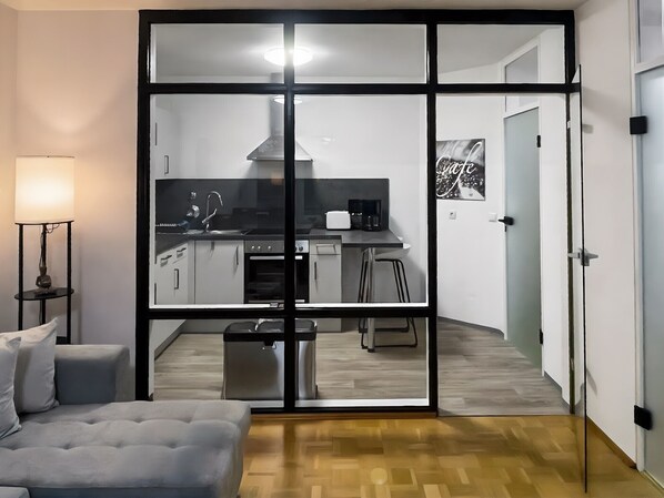 A modern living room with a sofa, a small table with a lamp, and a large sliding glass door leading to a kitchen with a stove, sink, and bar stools. - Apartment 'Bamberg Burgblick' with Balcony and Wi-Fi (Bamberg)