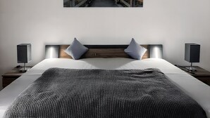 A bedroom with a wooden headboard, two bedside lamps, a large bed with a textured blanket, and a framed picture of a coastal scene.