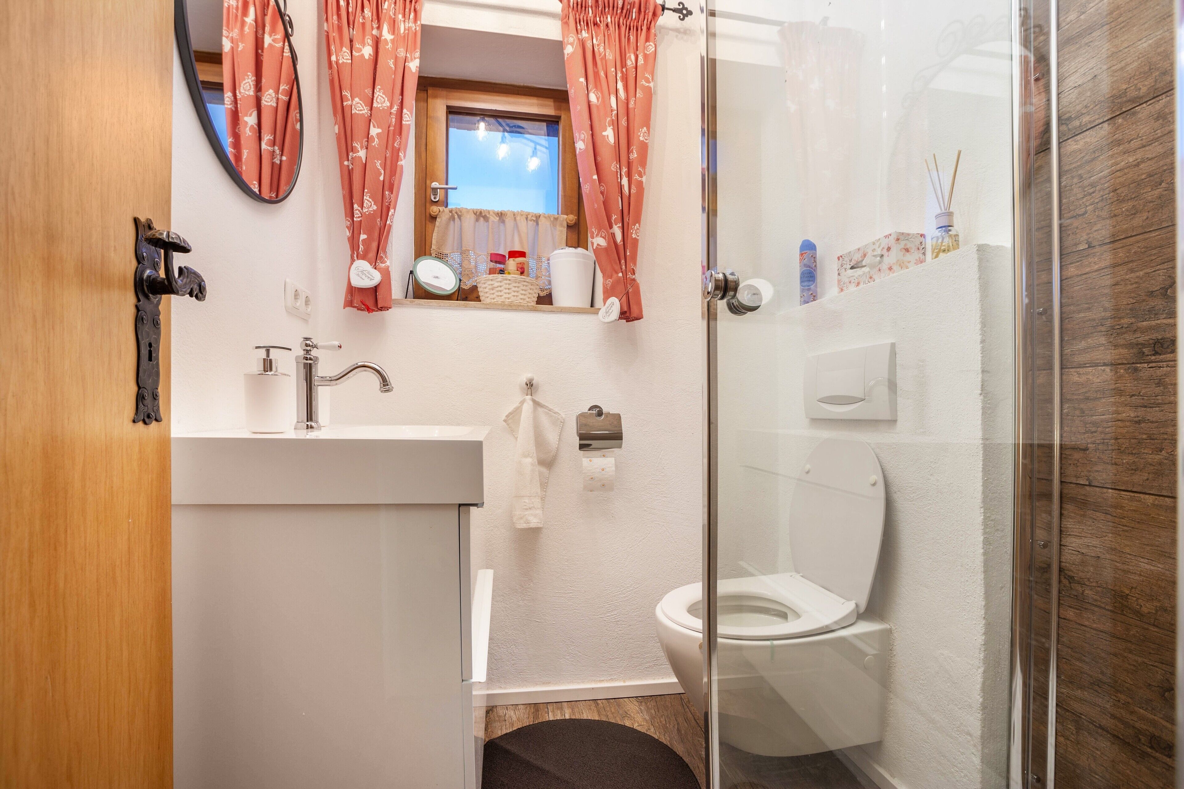 Een badkamer met een douchecabine van glas, toilet, wastafel met spiegel en raam met gordijnen.
