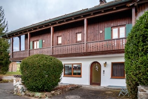 A wooden building with a balcony, a stone wall, and a well-maintained hedge. - Apartment 'In Urspring Am Lechstausee' with Mountain View, Shared Garden and Balcony (Steingaden - Urspring)