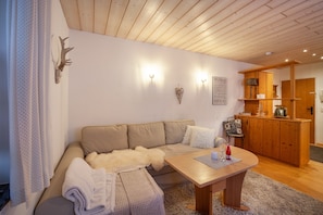 A cozy living room with a sofa, a wooden table, and a kitchen area in the background. - Apartment 'In Urspring Am Lechstausee' with Mountain View, Shared Garden and Balcony (Steingaden - Urspring)