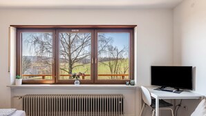 Une chambre avec une grande fenêtre, un radiateur, un lit, un bureau avec un ordinateur et une chaise.
