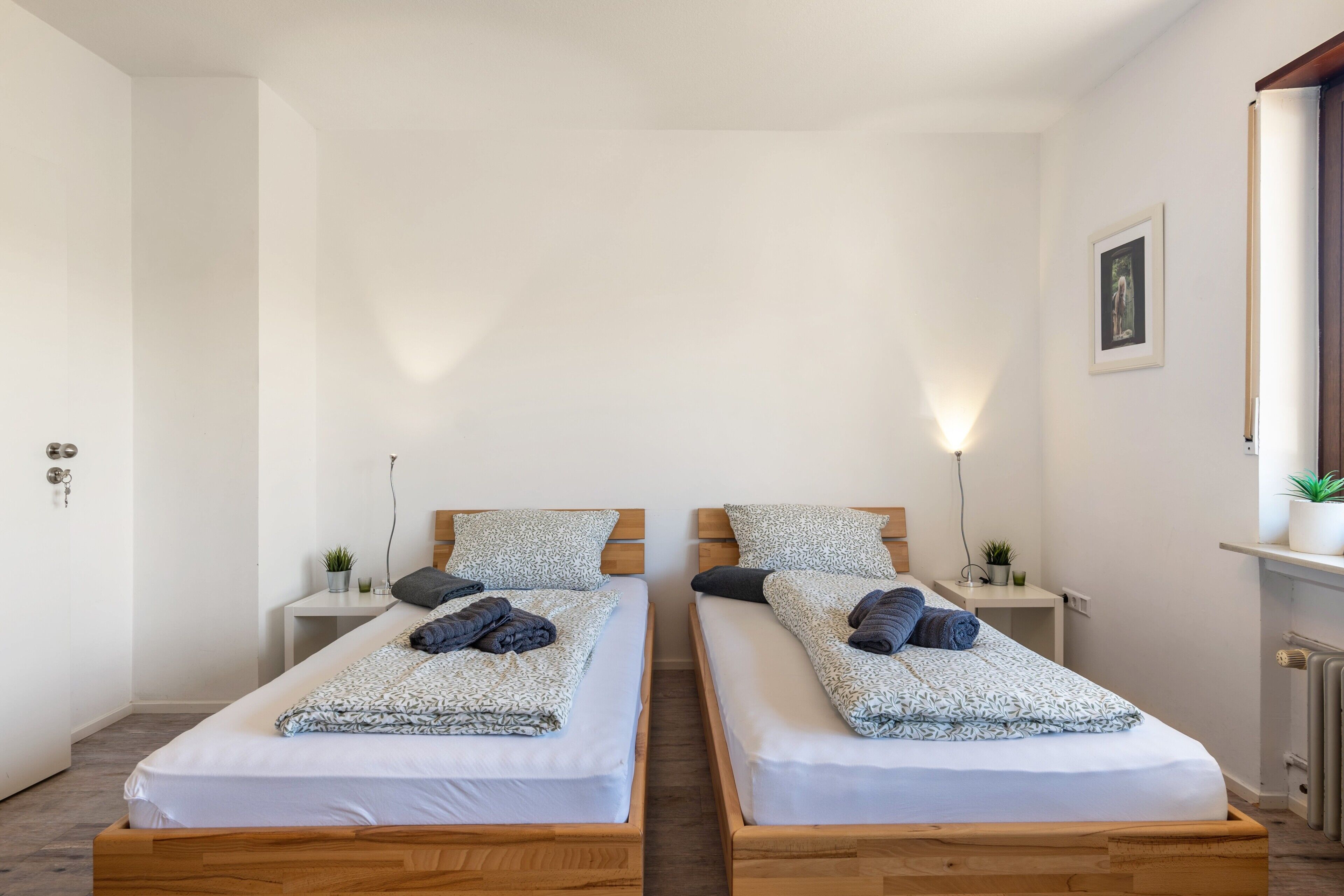 A double room with two beds, bedside tables, a wall-mounted light, and a small plant.