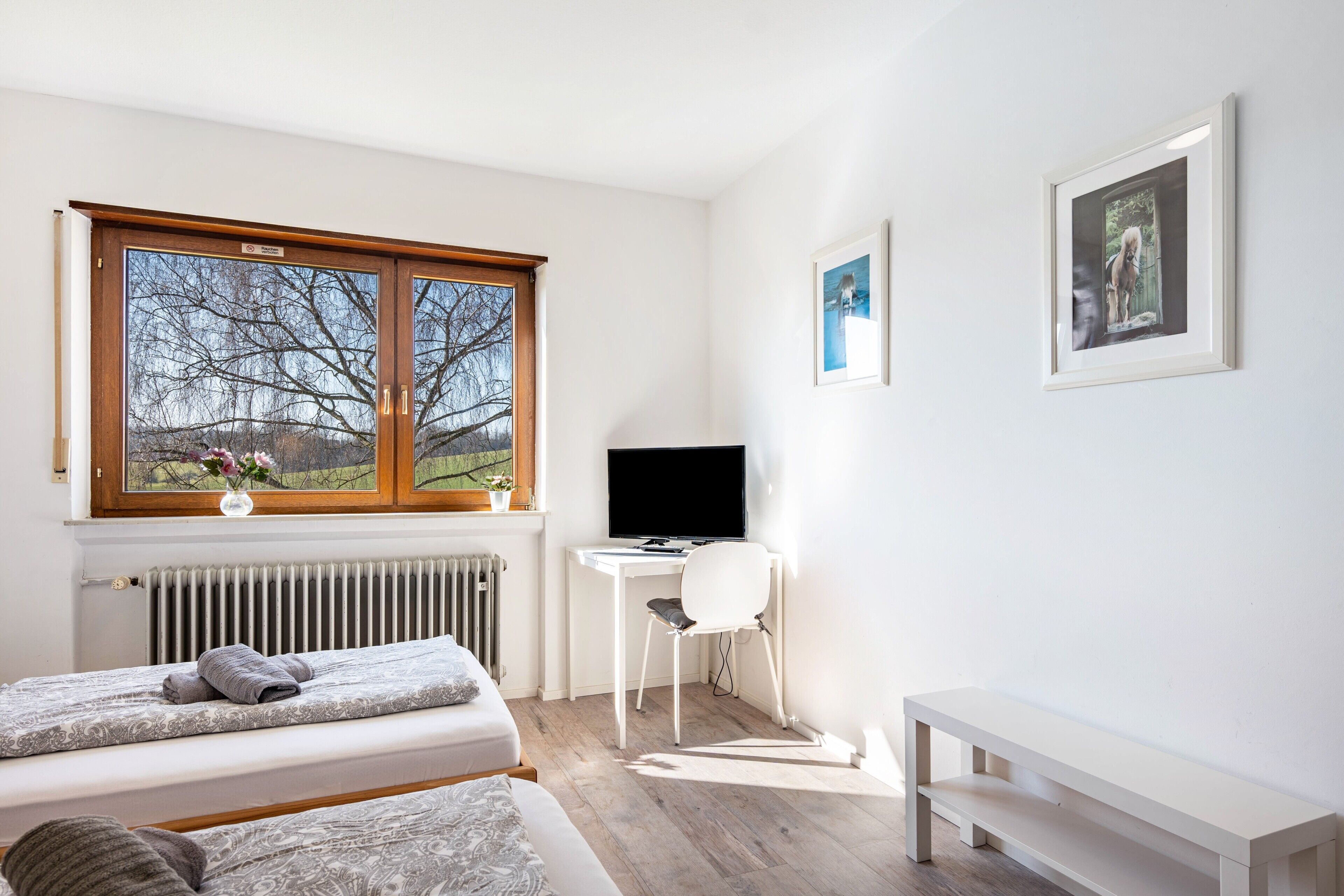Een slaapkamer met een bed, een bureau, een stoel, een bankje en een raam met uitzicht op bomen.