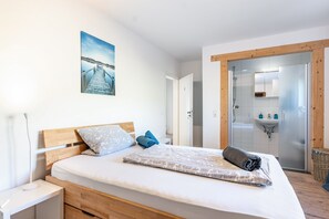 A bedroom with a wooden headboard, a bed with a patterned blanket, a nightstand, a wall-mounted lamp, a painting of a dock, and a bathroom with a glass shower door and sink. - Room 'Einzelzimmer' with Shared Terrace, Shared Garden and Wi-Fi (Dielheim)