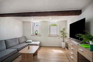 A modern living room with a sofa, coffee table, TV, and windows with plants.