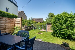 Une terrasse avec une table et des chaises, une balançoire et un jardin agrémenté d’une haie et d’un arbre.