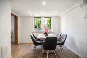 A dining area with a table set for four, surrounded by chairs, a window with a view of greenery, and a door leading to another room.