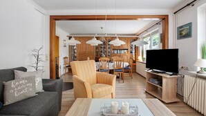A living room with a sofa, armchair, coffee table, TV, and dining area with chairs and a table. - Holiday Home 'Gerda' with Private Terrace, Private Garden and Wi-Fi (Stadum)
