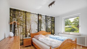 A bedroom with a wooden bed, a nightstand, a dresser, a window with a view of greenery, and a wall with a forest mural. - Holiday Home 'Gerda' with Private Terrace, Private Garden and Wi-Fi (Stadum)