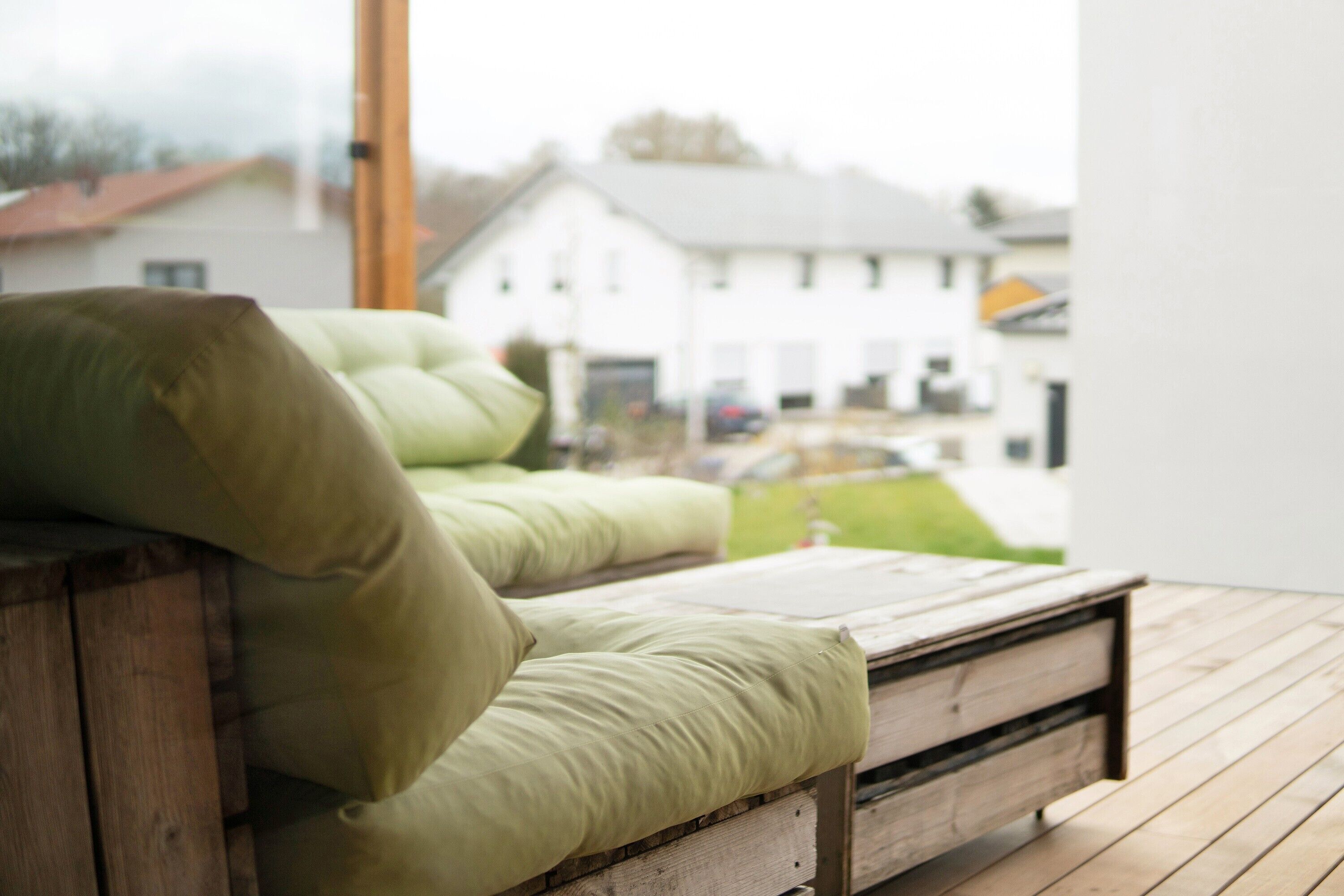 A wooden bench with green cushions, a wooden floor, and a view of a white house through a window.