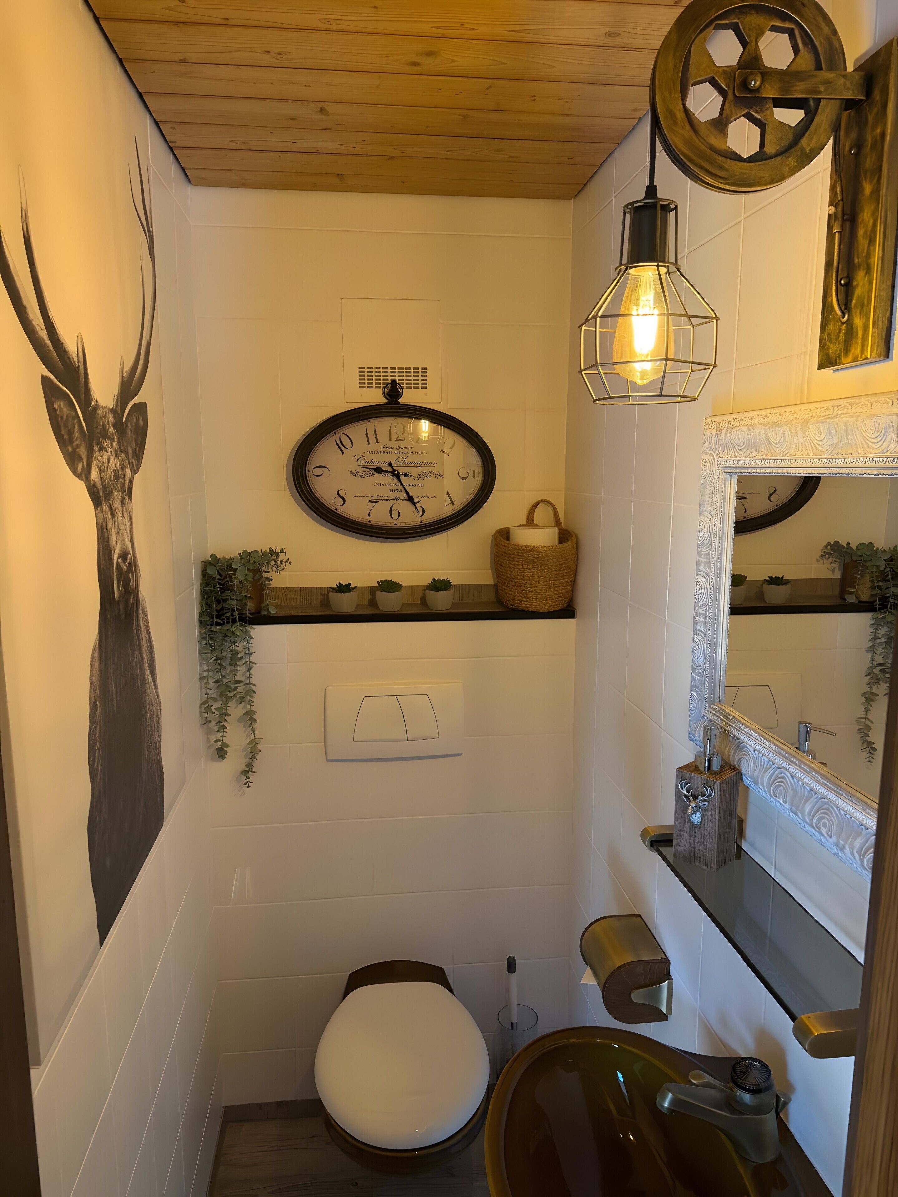 Ein Badezimmer mit Holzboden, einer Wanduhr, einer Toilette, einem Waschbecken mit Spiegel und einem dekorativen Hirschkopf.
