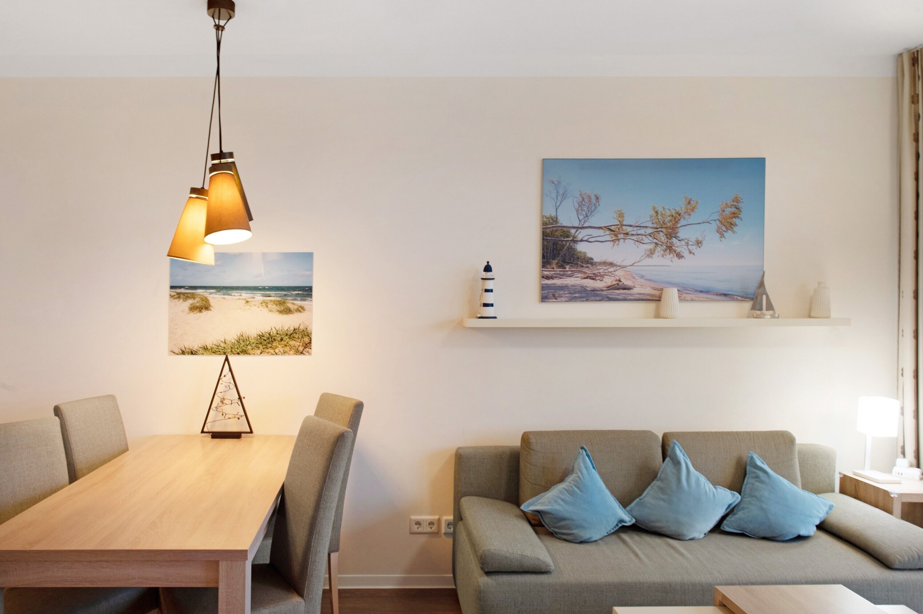 Ein Zimmer mit Esstisch aus Holz, zwei Stühlen, einem grauen Sofa mit blauen Kissen, einem an der Wand befestigten Regal mit Dekoration und zwei gerahmten Bildern.