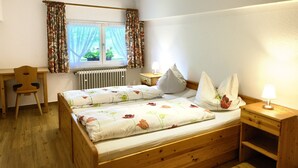 A room with a wooden bed, a desk, a chair, a window with curtains, and a bedside lamp.
