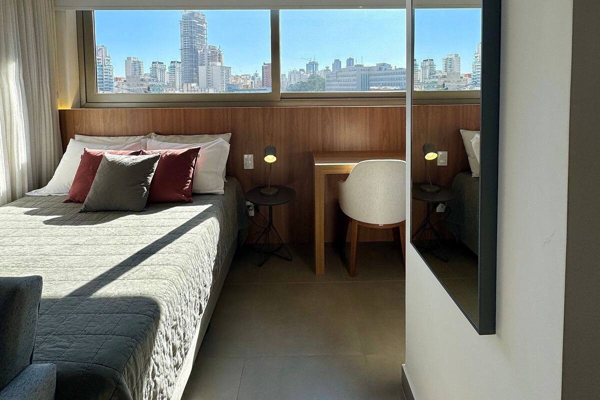 Ett modernt hotellrum med en stor säng, ett skrivbord, en stol och utsikt över stadens silhuett genom stora fönster.