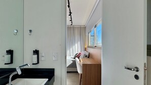 A modern bathroom with a sink, mirror, and towel rack. A bedroom with a bed and a desk is visible through an open door. - Apartamentos Prime-Prox a Oscar Freire (São Paulo)
