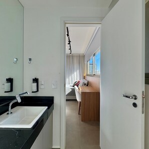 Un baño moderno con lavamanos, espejo y toallero. Se ve un dormitorio con cama y escritorio a través de una puerta abierta.