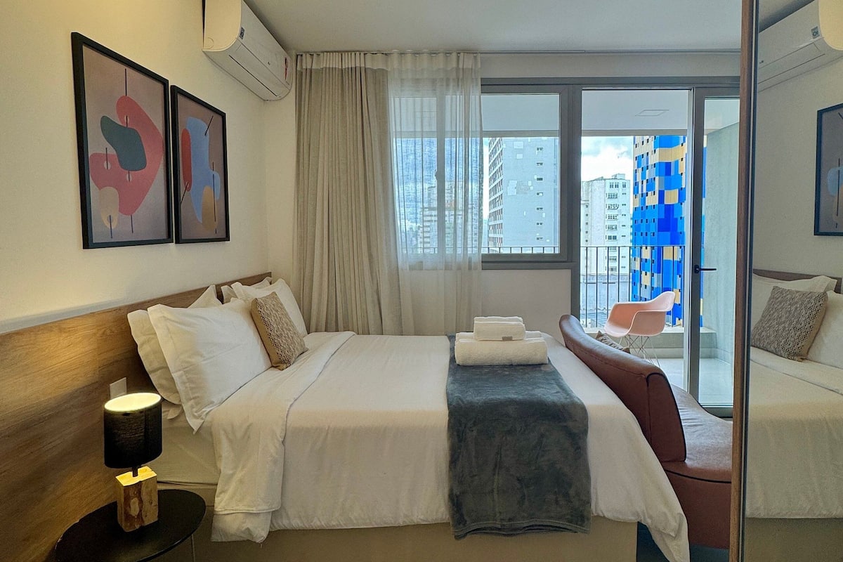 Quarto de hotel com cama, mesa de cabeceira, espelho e vista para a cidade pela janela.
