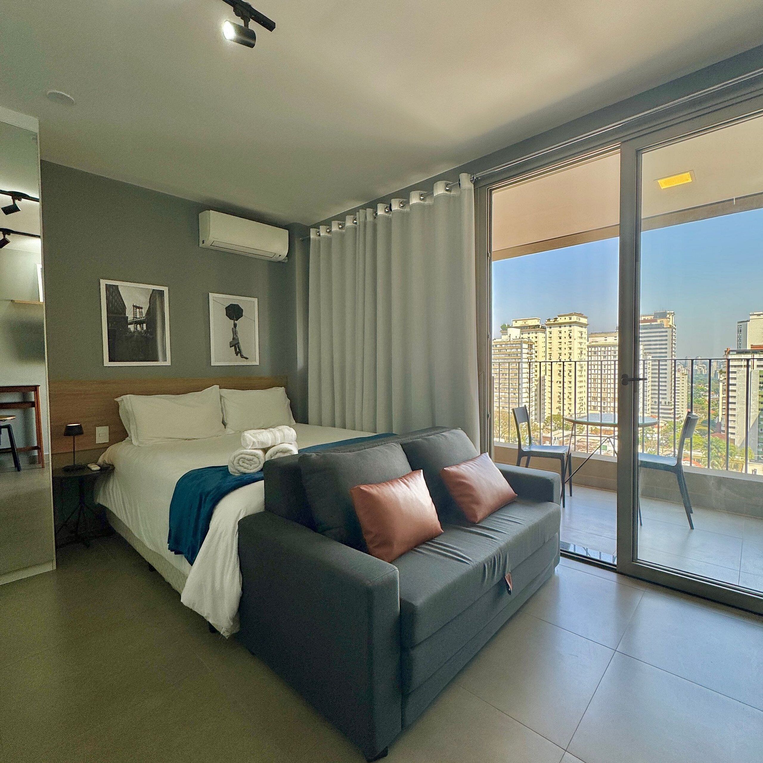 A modern hotel room with a bed, a sofa, a balcony with a city view, and a ceiling-mounted air conditioner.