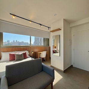 A modern hotel room with a large bed, a sofa, a desk, and a city view through the window.