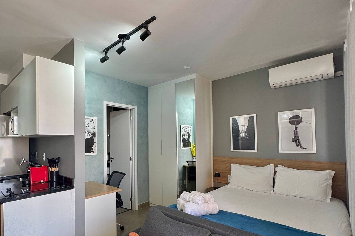 Quarto de hotel moderno com cozinha compacta, cama com roupa de cama branca, sofá cinza com almofadas laranja e obras de arte nas paredes.