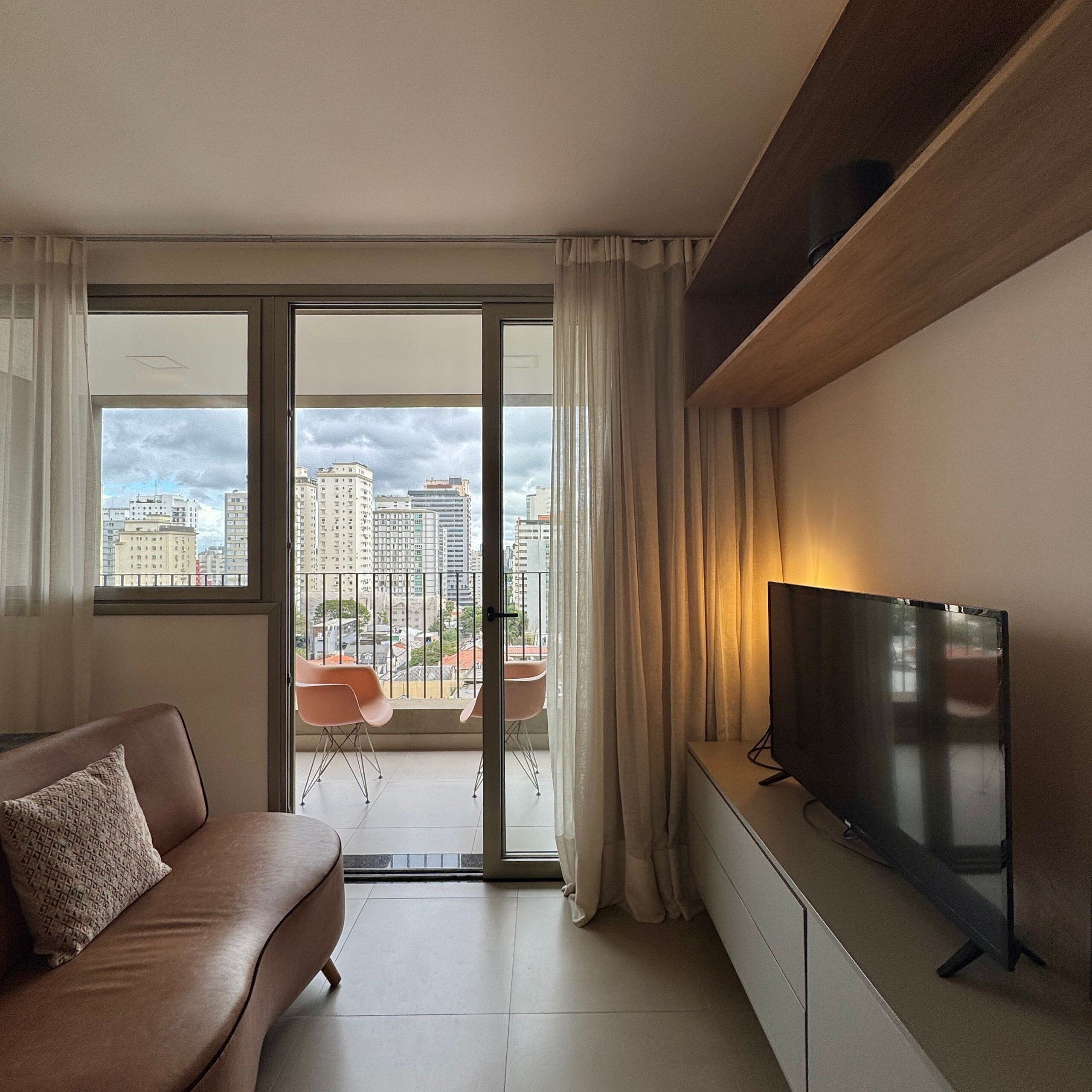 A modern living room with a sofa, a flat-screen TV, and a sliding glass door leading to a balcony with a city view.