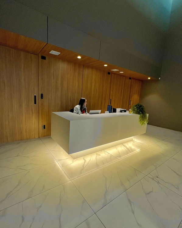 A reception desk with a woman seated behind it, a potted plant to the right, and wooden paneling on the walls. - Apartamentos Prime-Prox a Oscar Freire (São Paulo)