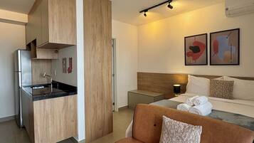 Ein kompaktes Hotelzimmer mit Küchenzeile, einem Bett mit Kopfteil, einem Nachttisch und zwei abstrakten Wandbildern.